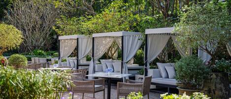 An outdoor seating area with cushioned chairs and tables, surrounded by lush greenery and trees.