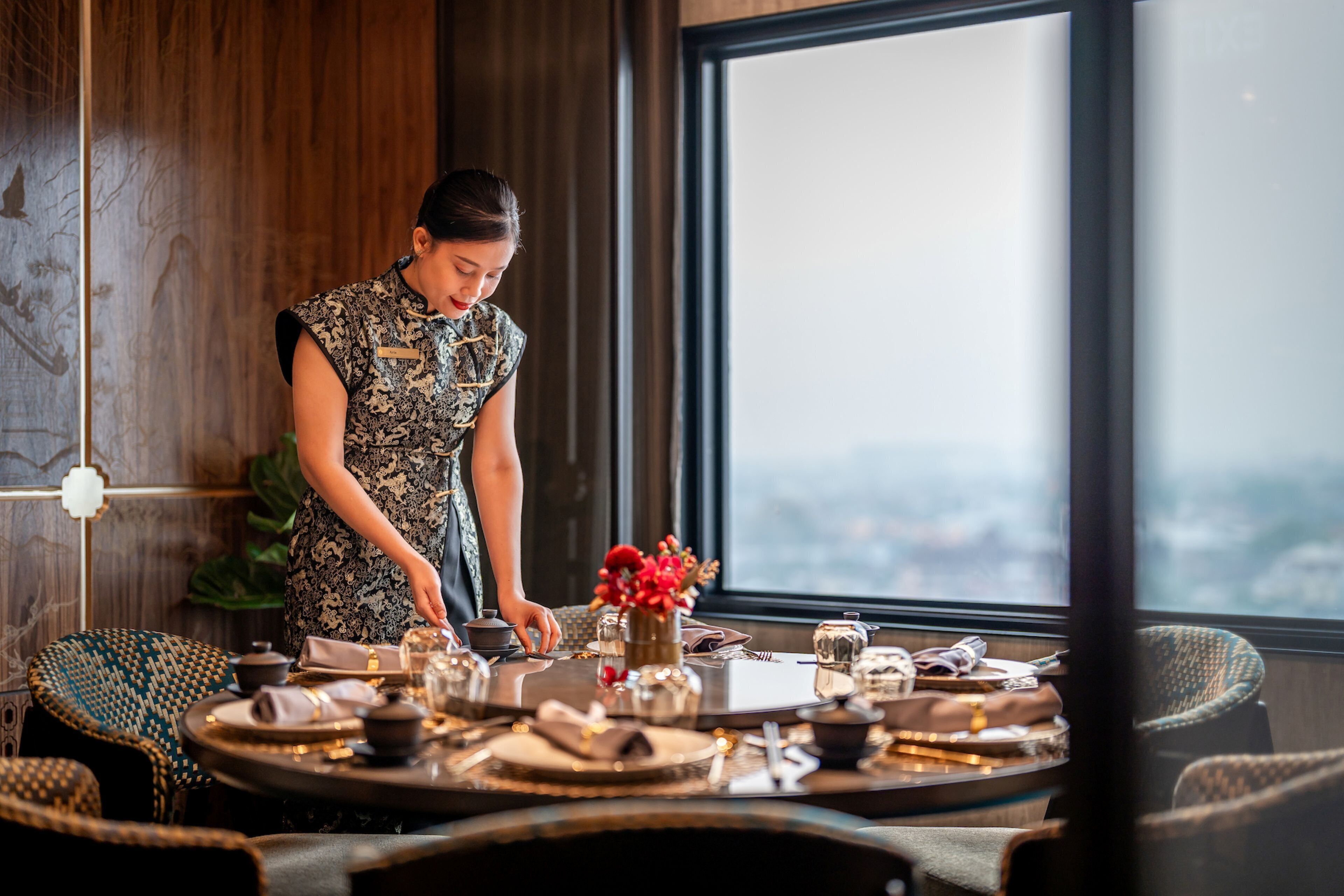 A dining area with a table set for two, a vase with red flowers, and a view of the city through a large window.