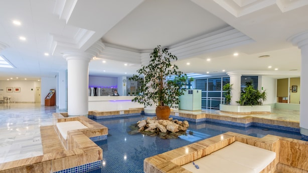 A spacious indoor pool area with a central island, seating areas, and a reception desk in the background. - Illot Suites (Capdepera)