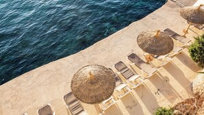 Vista aérea de una playa con aguas azules cristalinas, una orilla de arena y varias sombrillas de paja con sillones de descanso.