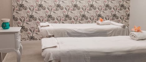 Een spa-ruimte met twee behandelbedden, een klein tafeltje met een vaas, en bloemenbehang.