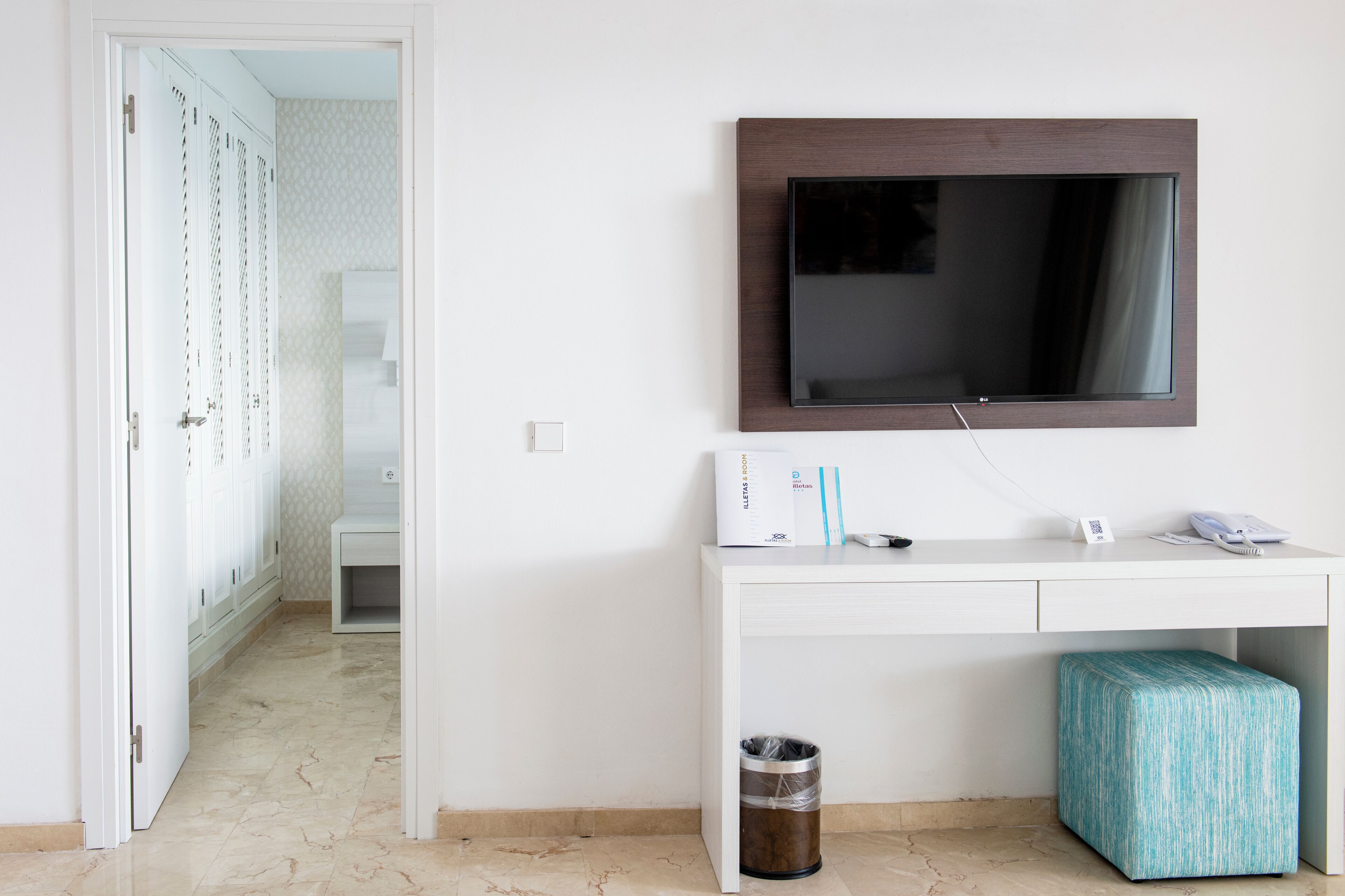Een hotelkamer met een wit bureau, een wandtelevisie, een prullenbak en een gestreept voetenbankje.