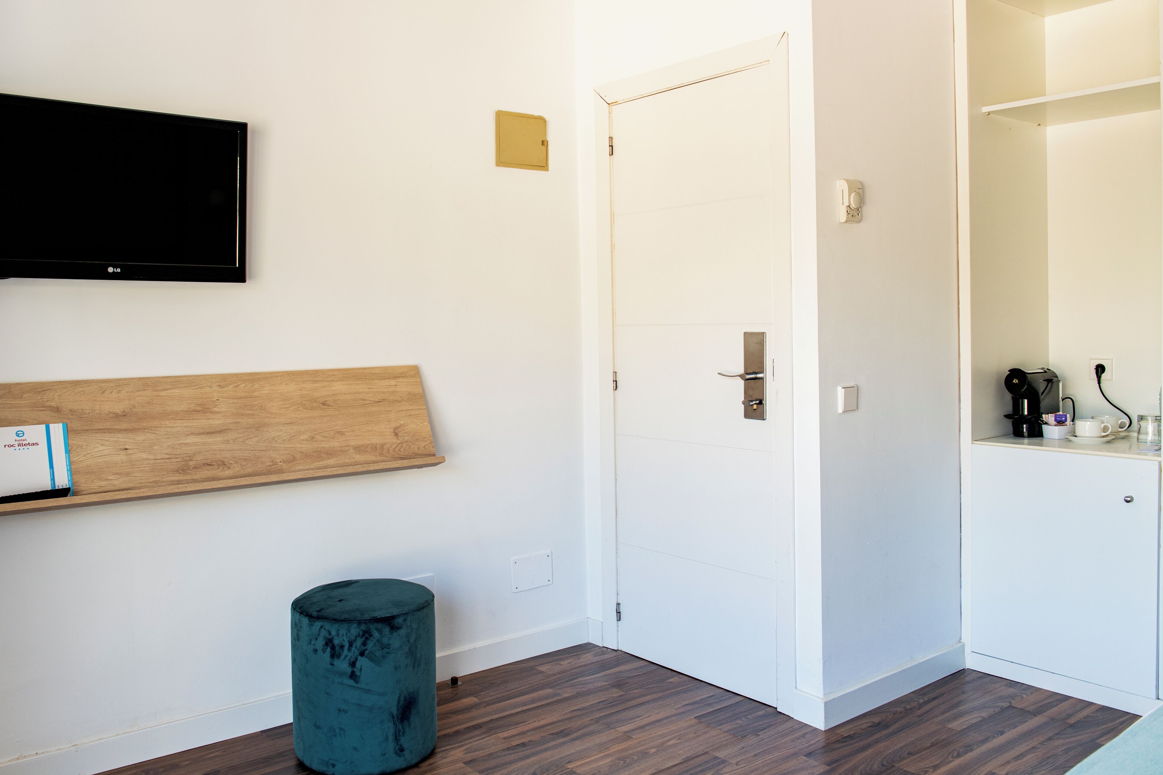 Une pièce avec une étagère en bois, un pouf turquoise, une porte blanche et un téléviseur à écran plat fixé au mur.