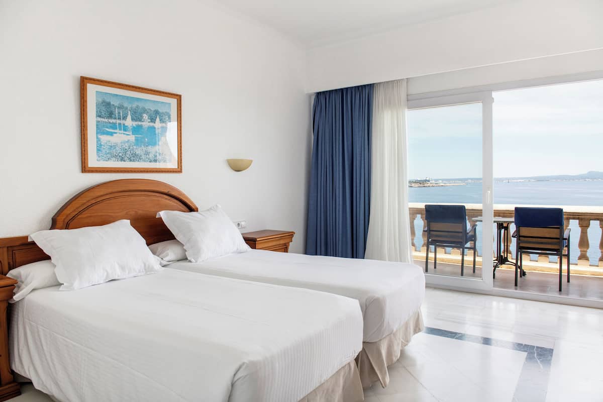 A hotel room with two beds, a balcony with chairs, a framed picture on the wall, and a view of the sea.