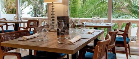 A dining area with wooden tables and chairs, a lamp, and a view of palm trees through large windows.