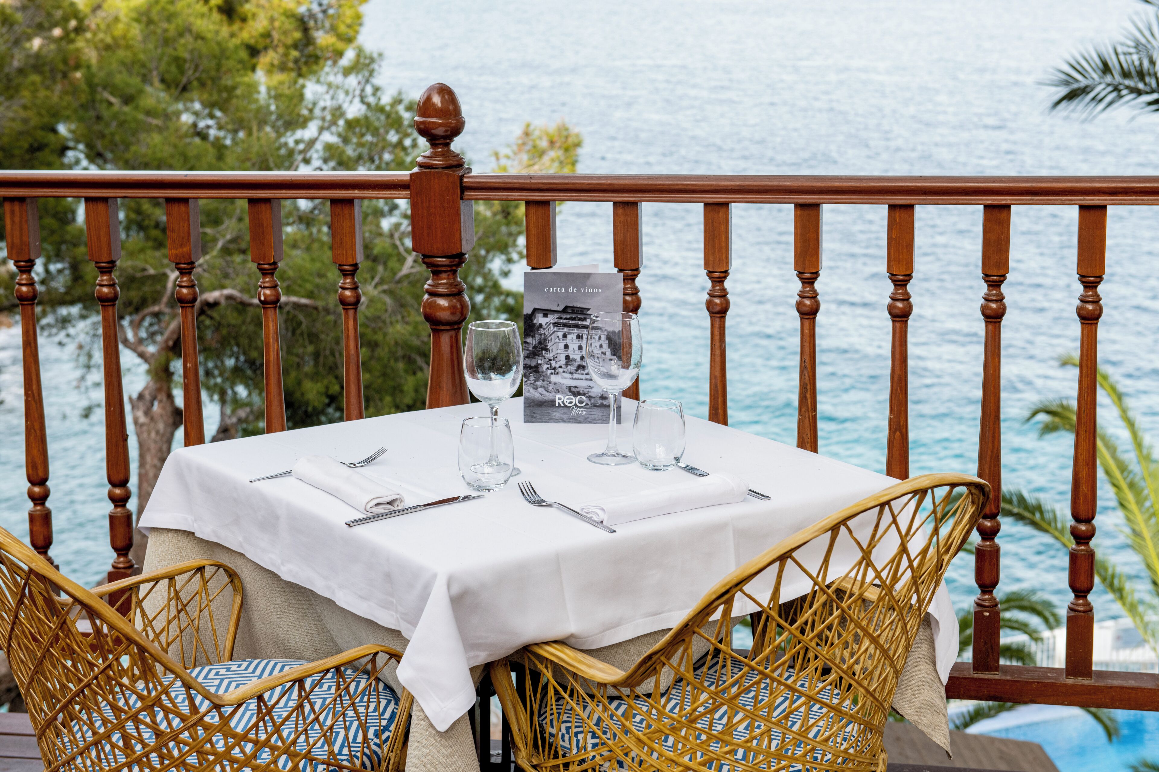 Mesa para dos con mantel blanco, dos copas de vino y una carta en un balcón con vista al mar.