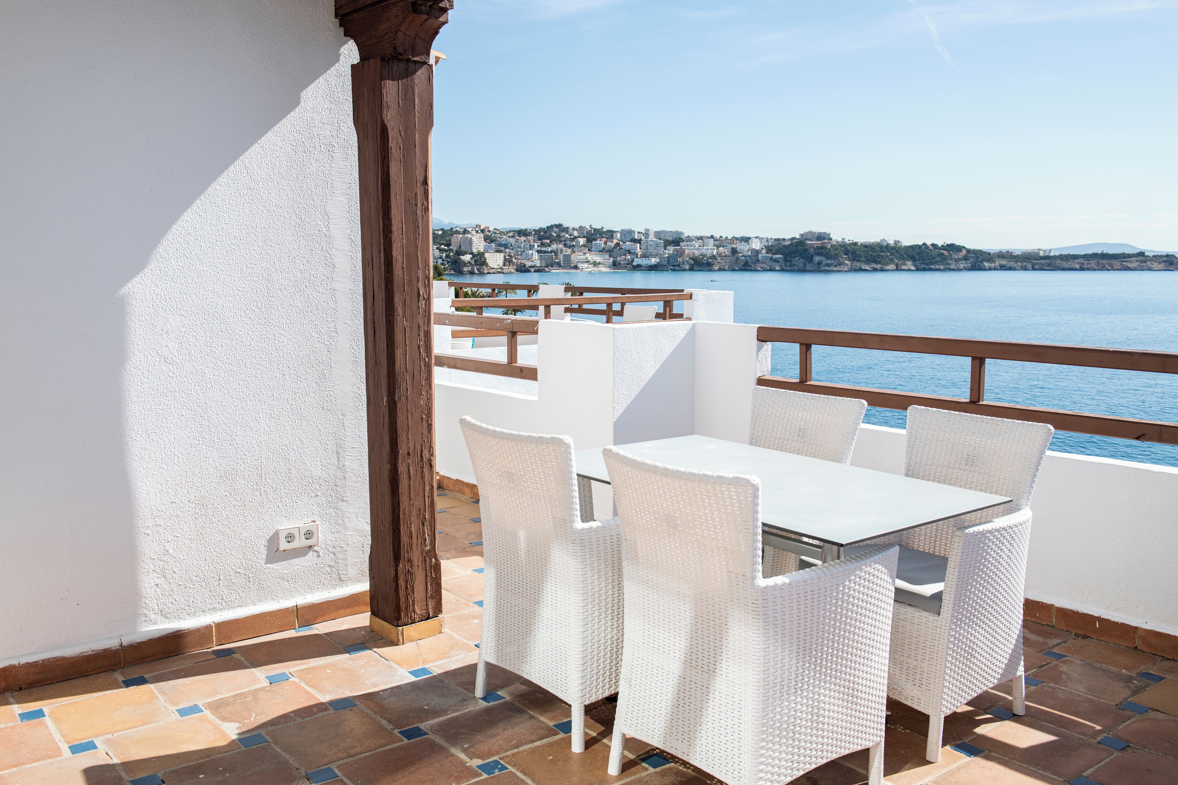 A balcony with white wicker furniture, a tiled floor, and a view of a coastal town.