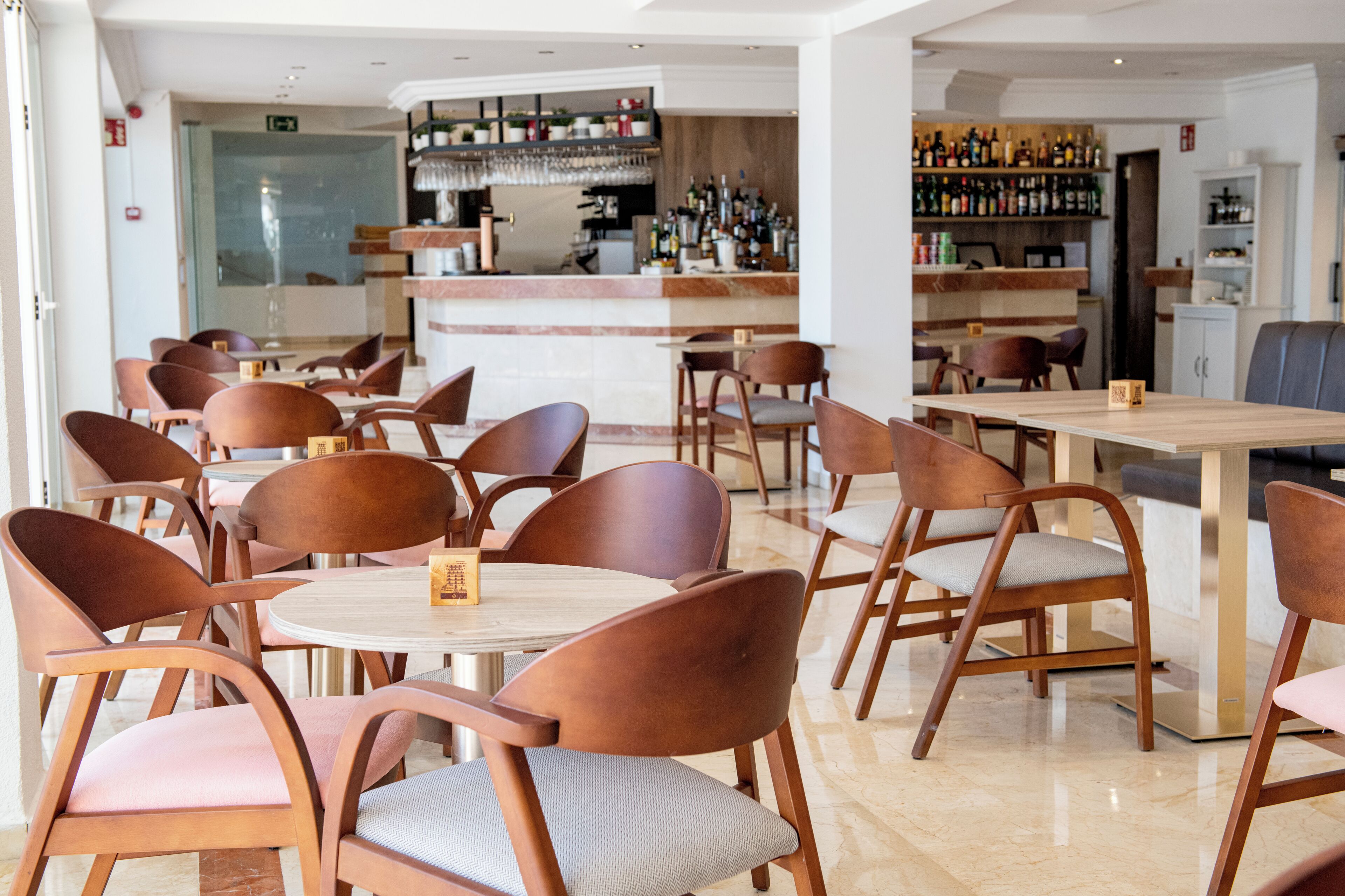 Un restaurant moderne avec des tables et des chaises en bois, un bar doté d’étagères remplies de bouteilles, et un intérieur propre et bien éclairé.