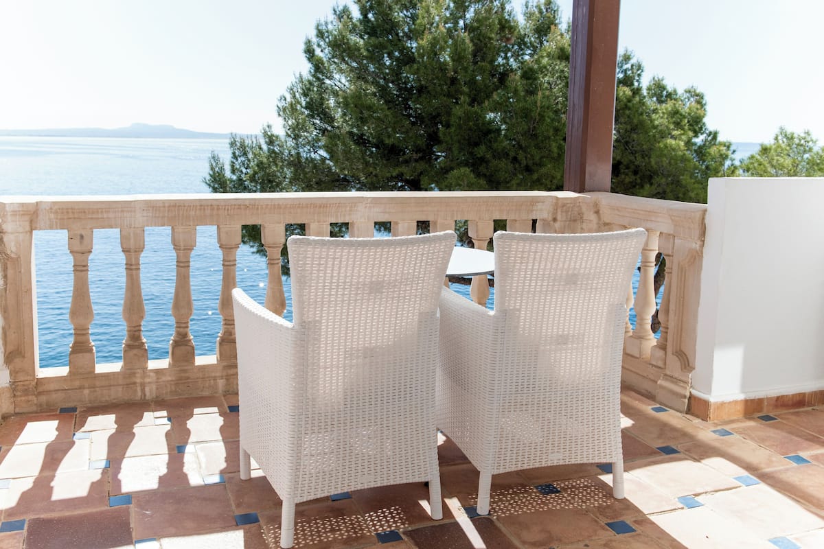 A balcony with two white wicker chairs and a small table, overlooking a body of water and a mountainous landscape.