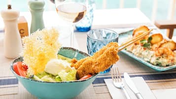 Une table à manger avec un saladier bleu contenant une salade, un plat frit et un verre de vin blanc.