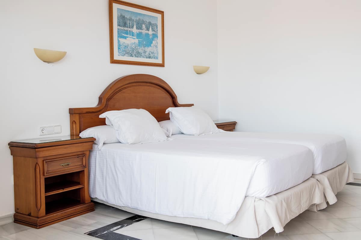 A double bed with white bedding, a wooden nightstand with a drawer, and a wall-mounted light fixture.