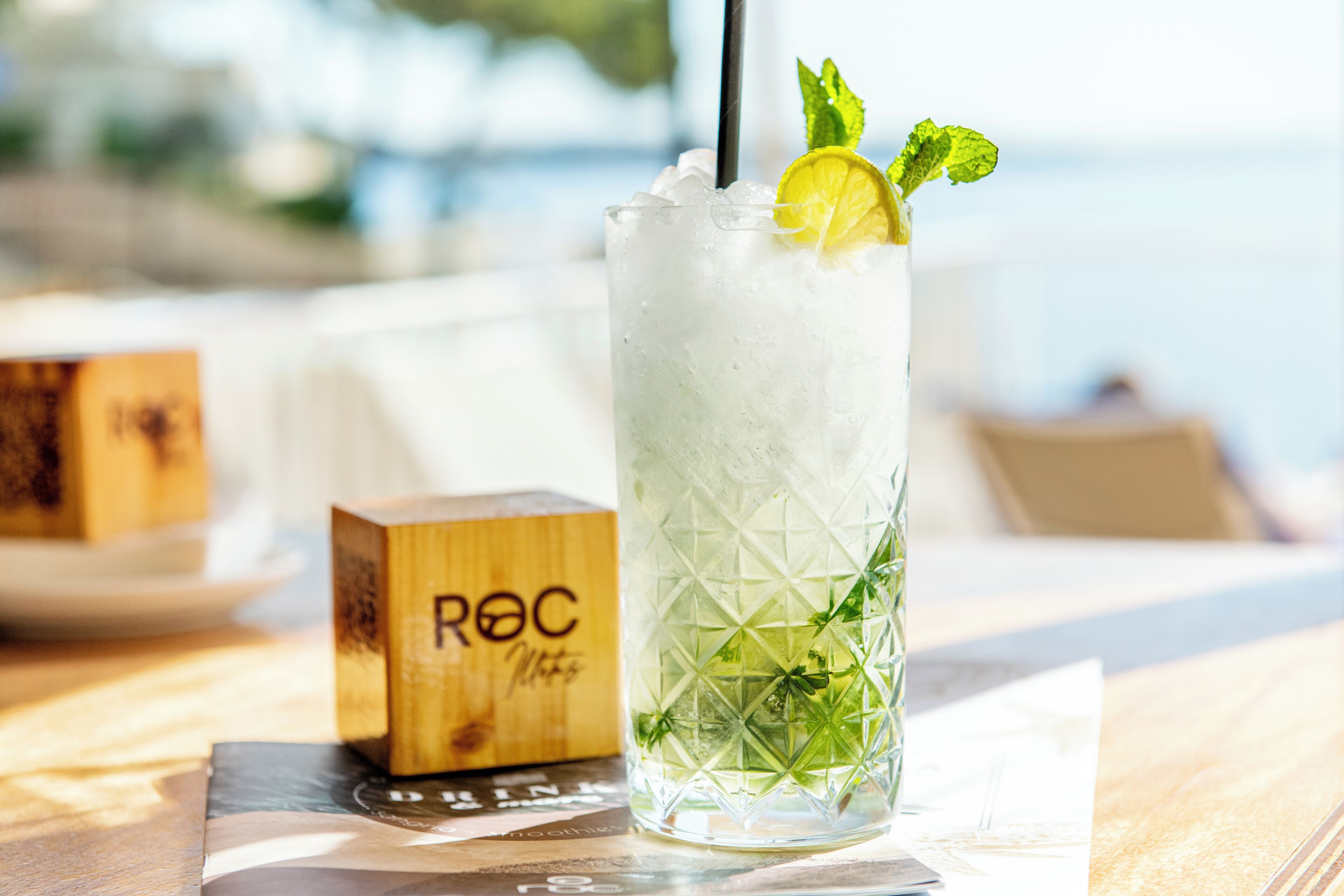 A glass of iced drink with mint and a lime slice, placed on a wooden table with a wooden block labeled "ROC" in the background.