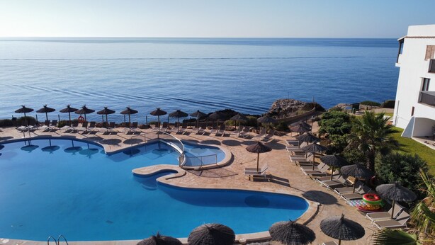 Zona de piscina con múltiples tumbonas y sombrillas, rodeada por un edificio con vistas al mar.