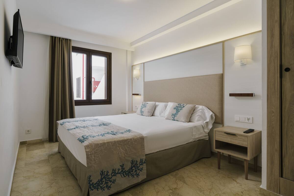 A hotel room with a bed, bedside tables, a TV, and a window with curtains.