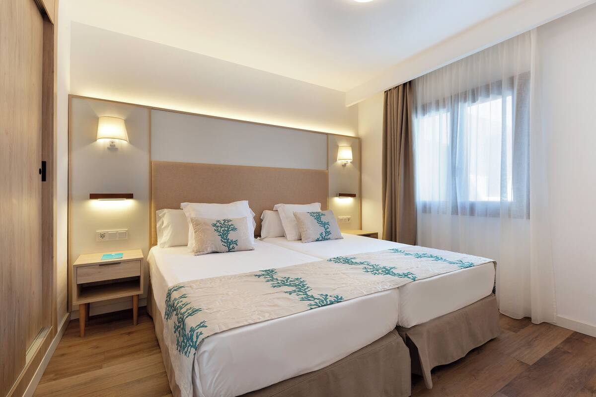 A double bed with a beige and white bedspread, two bedside tables with lamps, a wooden nightstand, and a window with sheer curtains.