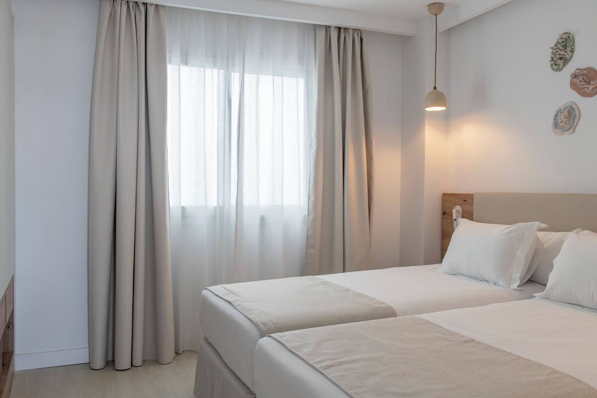 A hotel room with two beds, a window with sheer curtains, a wall-mounted light fixture, and a wooden headboard.