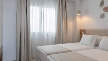 Habitación de hotel con dos camas, una ventana con cortinas traslúcidas, una lámpara fijada a la pared y un cabecero de madera.