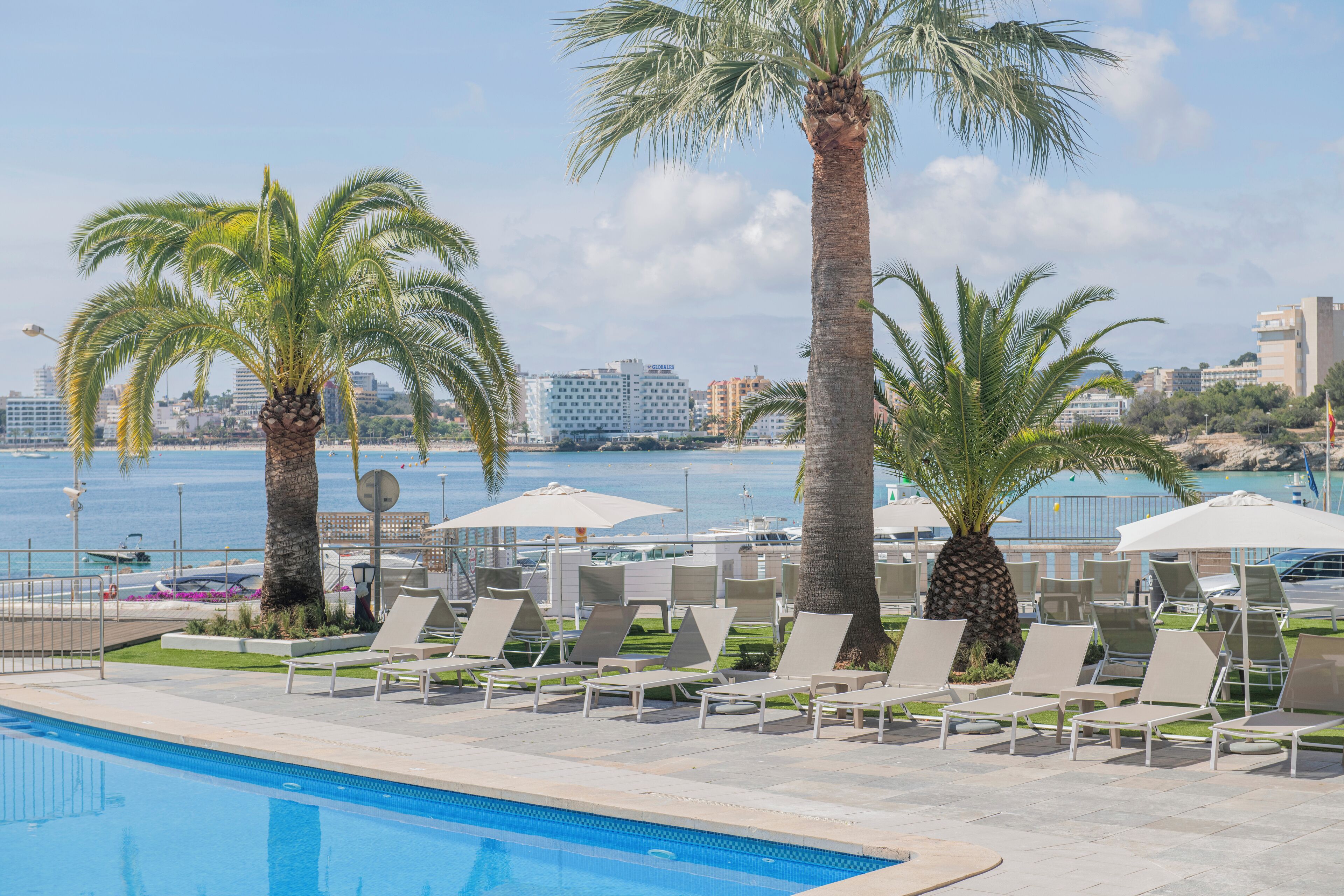 Ein Poolbereich mit Liegestühlen und Sonnenschirmen, Palmen und im Hintergrund Meer und Gebäude.
