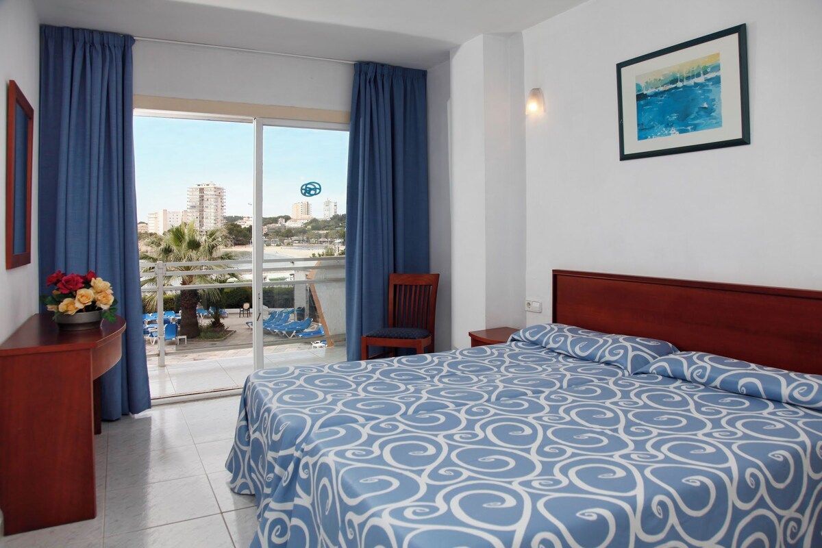 A hotel room with a large bed, a wooden nightstand, a chair, and a view of the city through a glass door.