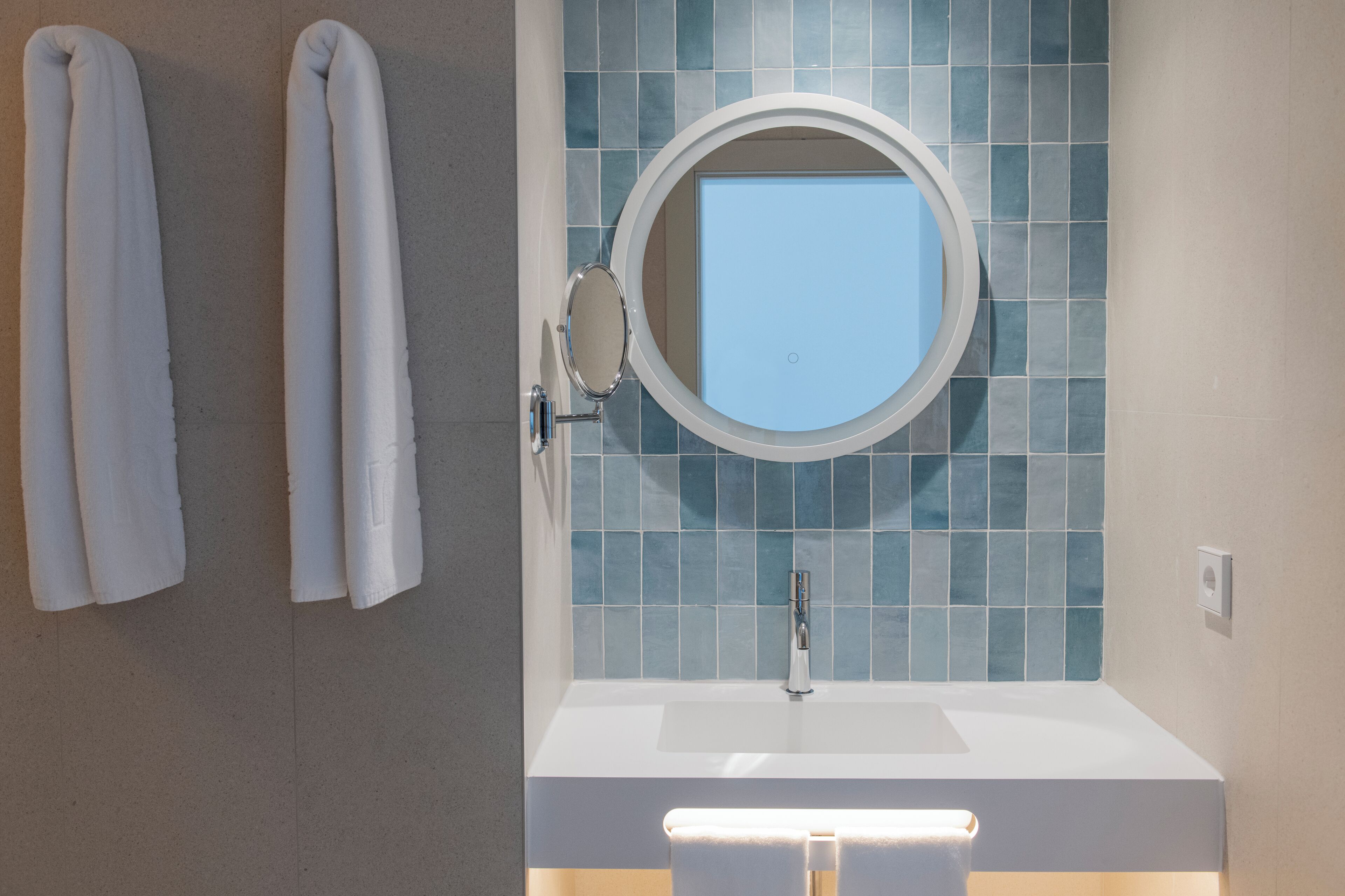 Un bagno con un lavandino bianco, uno specchio rotondo e una parete rivestita di piastrelle blu.