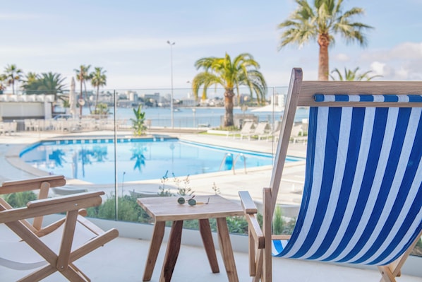 A wooden deck with a striped beach chair, a small table with sunglasses, and a view of a swimming pool and palm trees.