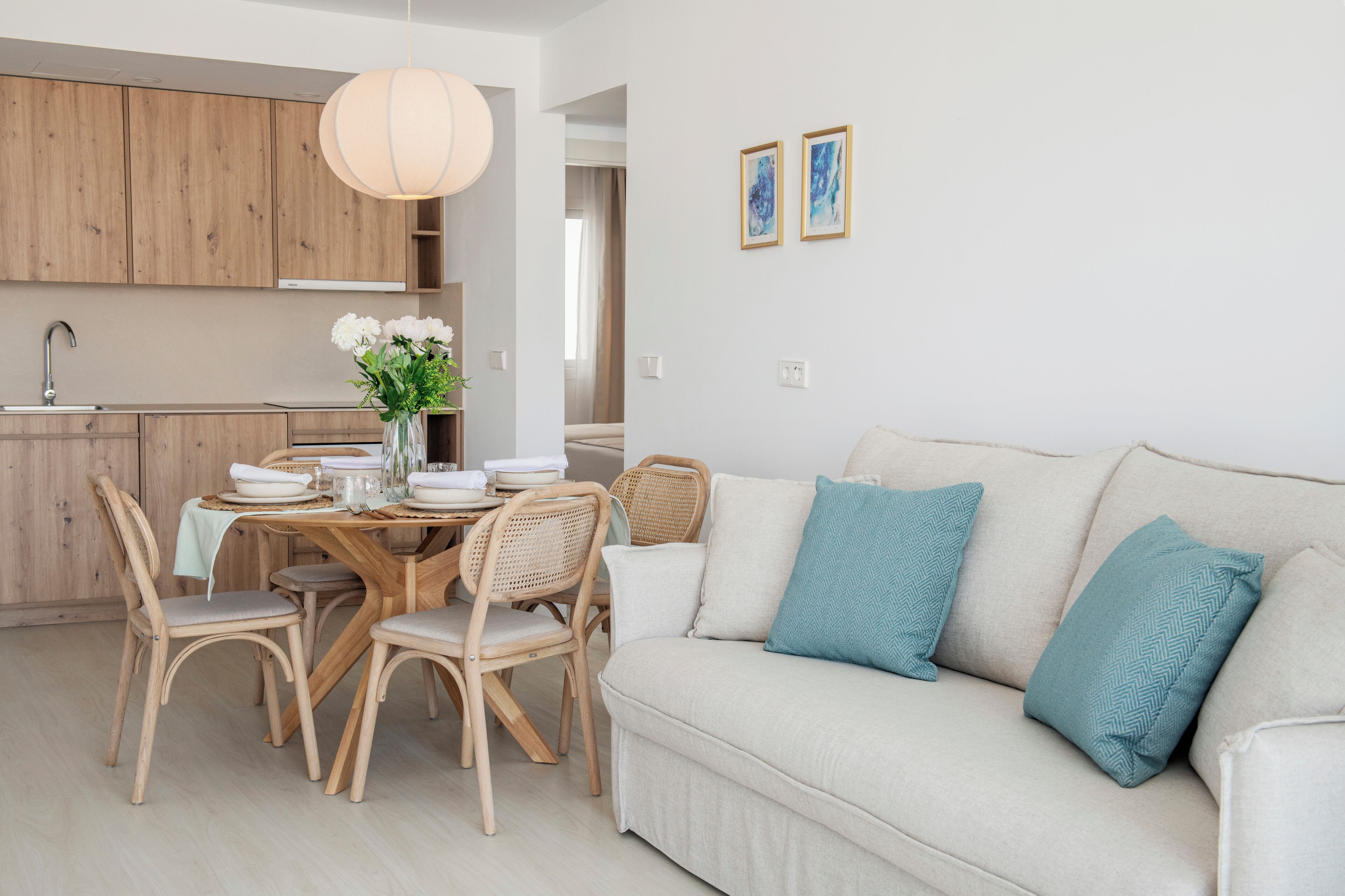 Una cocina moderna con armarios de madera, una mesa de comedor con platos blancos y un jarrón de flores, y un sofá de color claro con almohadones turquesa.