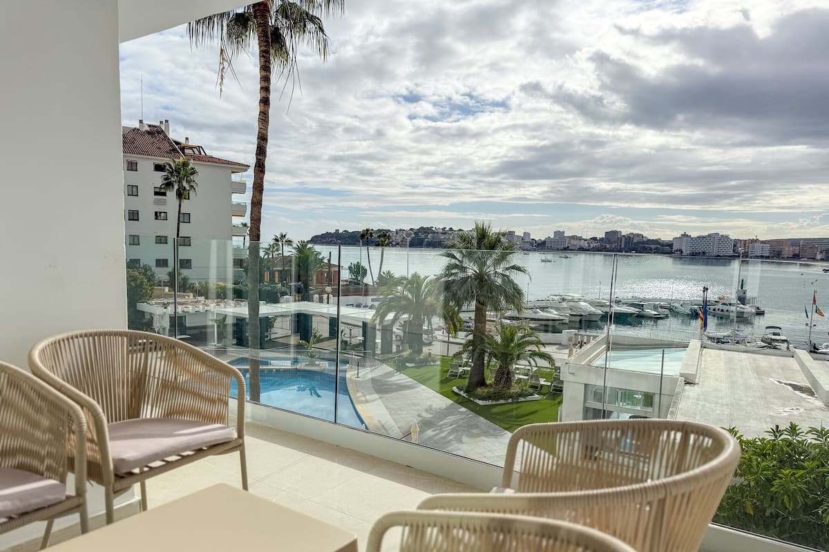 A balcony with wicker chairs and a table, overlooking a marina with boats and a cityscape.