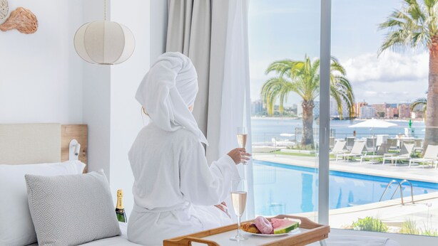 A person in a white robe sitting by a window with a view of a pool and palm trees. - Suites ROC Portonova - Newly Renovated 2026 (Calvia)
