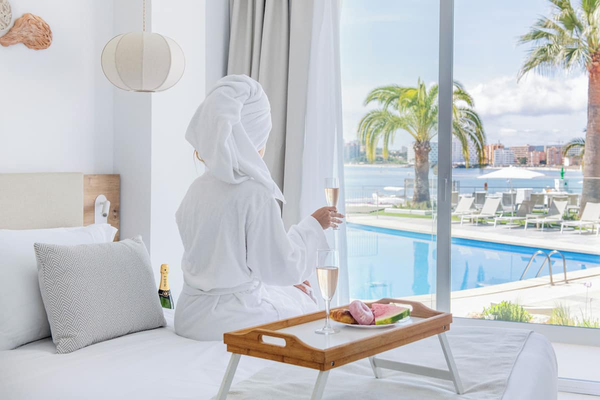 A person in a white robe sitting by a window with a view of a pool and palm trees.