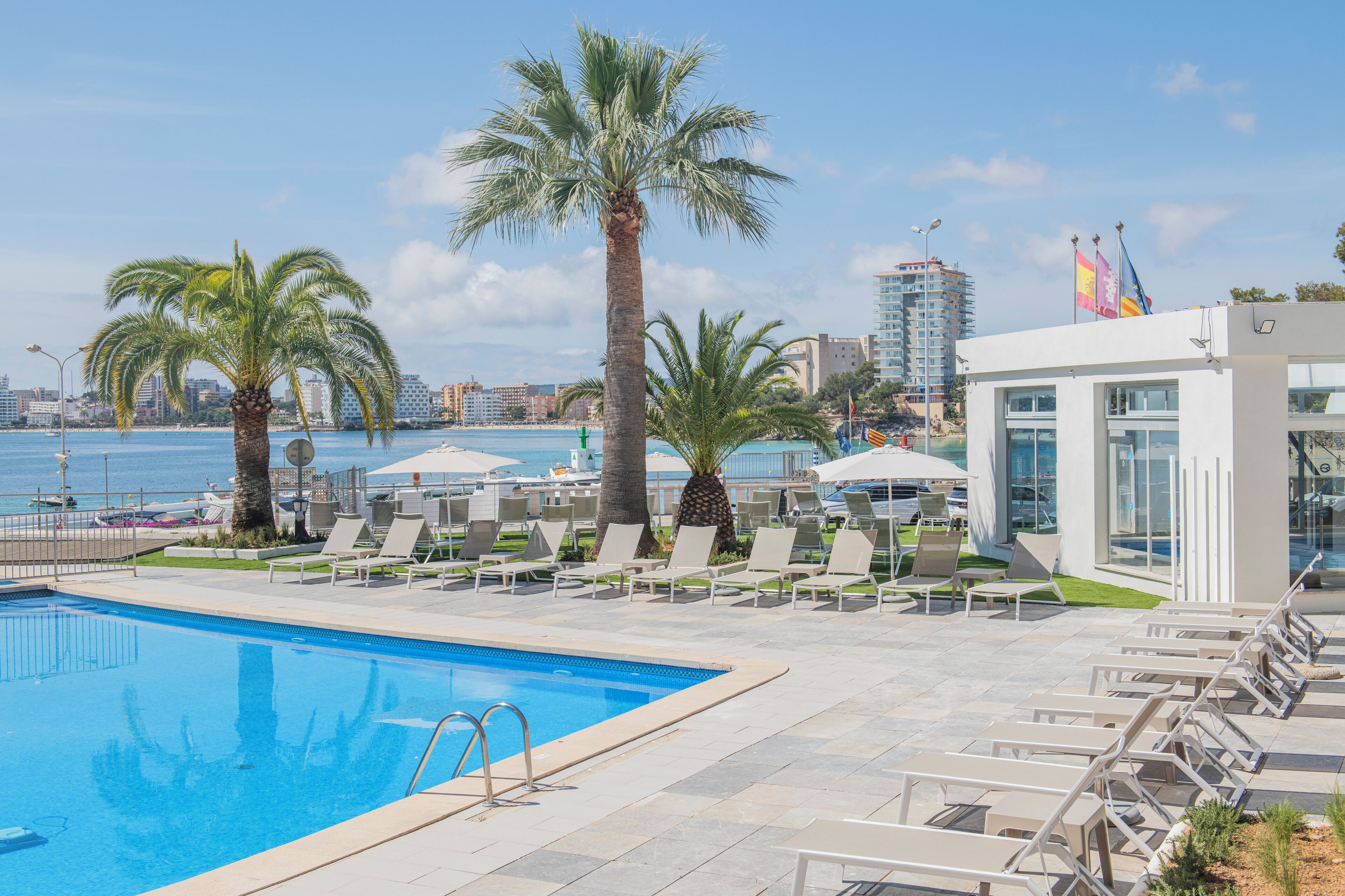 A pool area with lounge chairs and palm trees, overlooking a body of water and buildings.