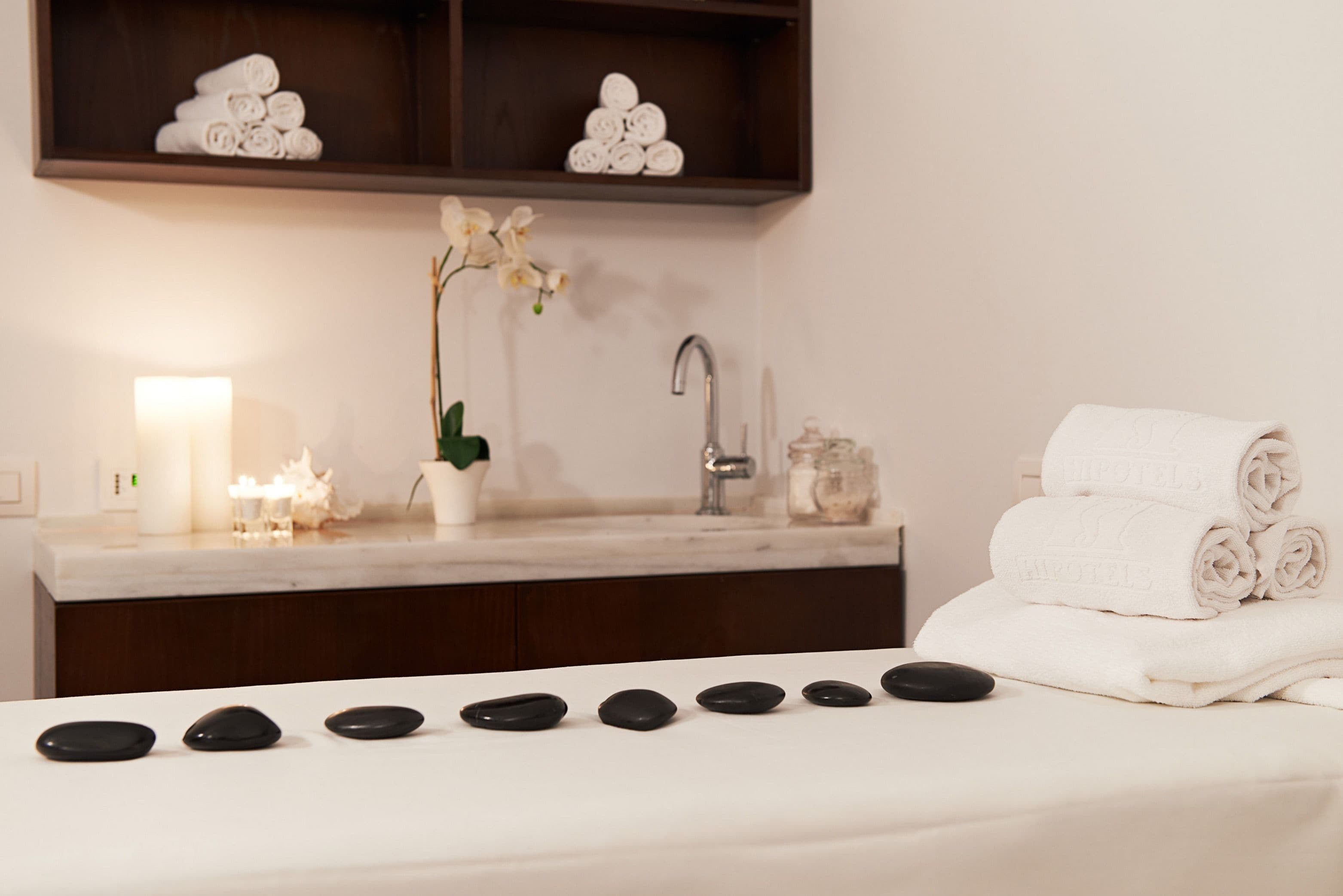 A spa room with a massage table, black stones, a sink, a potted orchid, and neatly stacked towels.