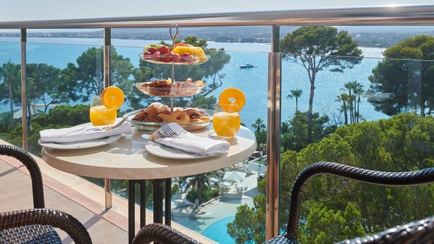 Desayuno con una bandeja de frutas, croissants y zumo de naranja en un balcón con vistas a una piscina y árboles.