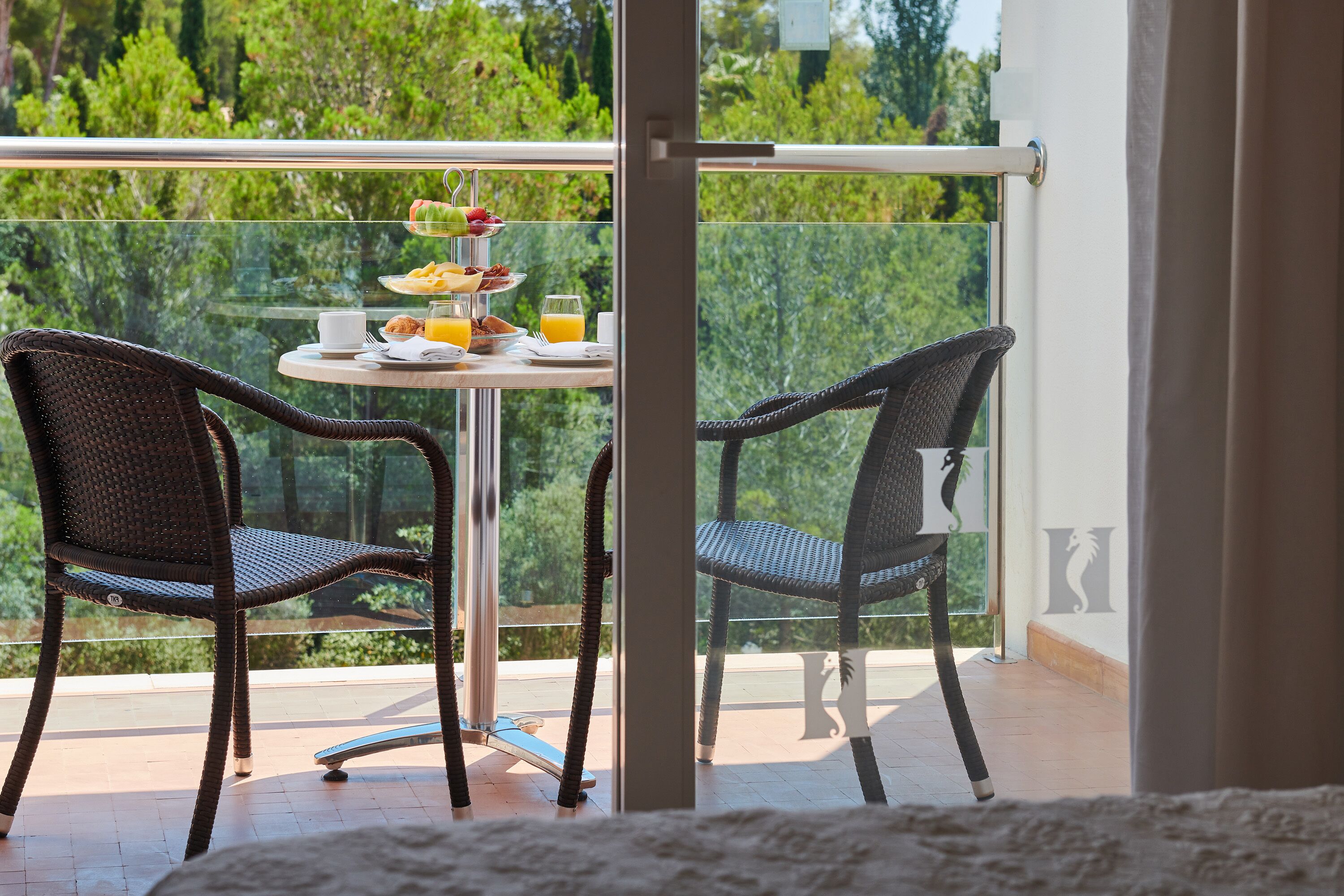 A balcony with wicker chairs, a table with breakfast items, and a glass railing overlooking greenery.
