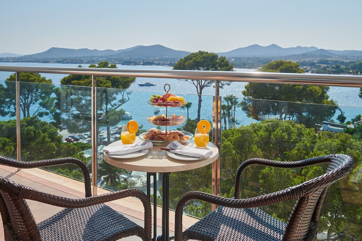 Un balcon avec des fauteuils en osier, une table où sont disposés des éléments pour le petit-déjeuner, et une vue panoramique sur un lac et des montagnes.
