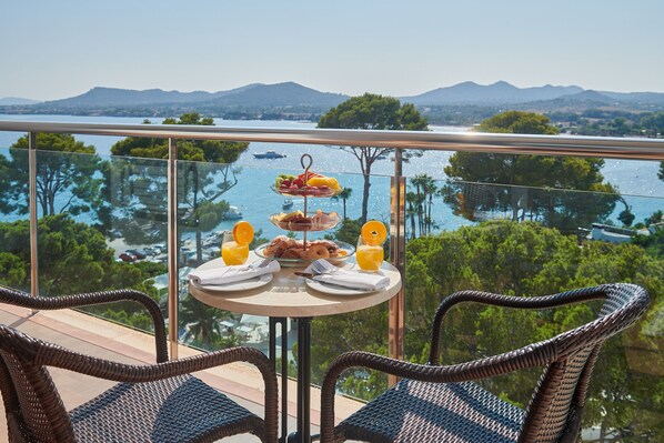 Un balcon avec des fauteuils en osier, une table où sont disposés des éléments pour le petit-déjeuner, et une vue panoramique sur un lac et des montagnes.