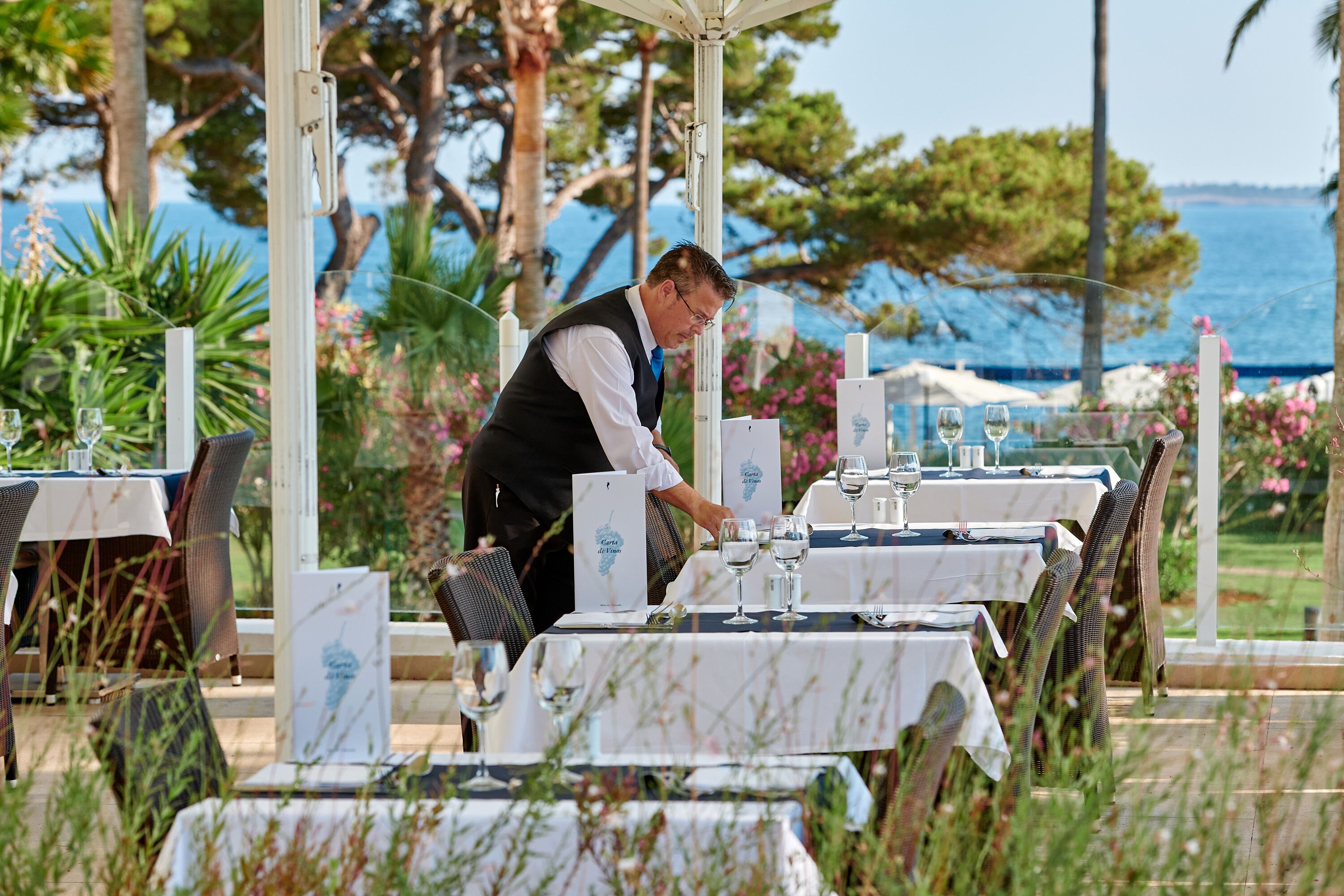 Un camarero preparando una mesa con manteles blancos y copas de vino en una zona de comedor al aire libre con vistas al mar.