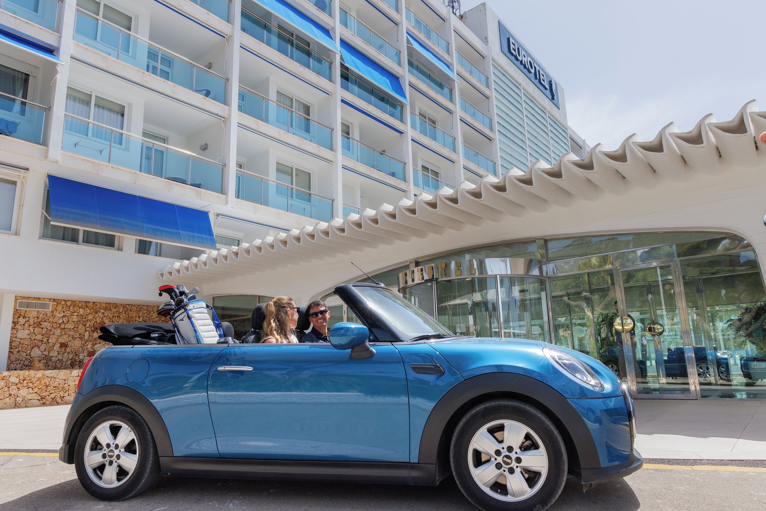Ein blauer Cabrio steht vor einem Hotel mit modernem Design und einem Schild, auf dem 'EUROTEI' steht.