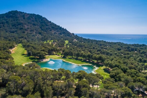 An aerial view of a golf course with a lake, surrounded by dense forest and a mountain in the background. - Hipotels Eurotel Punta Rotja & Spa (Son Servera)