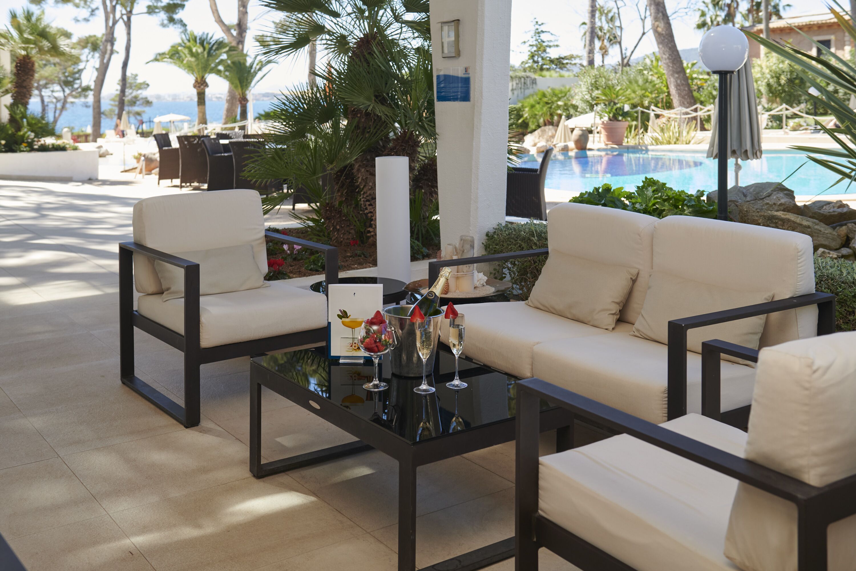 A poolside lounge area with a sofa and chairs, a glass table with drinks, and a view of the pool and palm trees.