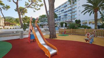 A playground with a slide and swings, surrounded by palm trees and a building with balconies.