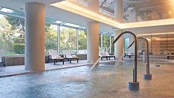 A modern indoor pool area with large columns, seating, and a view of greenery outside.