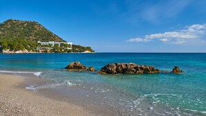 Una playa con agua turquesa cristalina, afloramientos rocosos y una colina con edificios al fondo.
