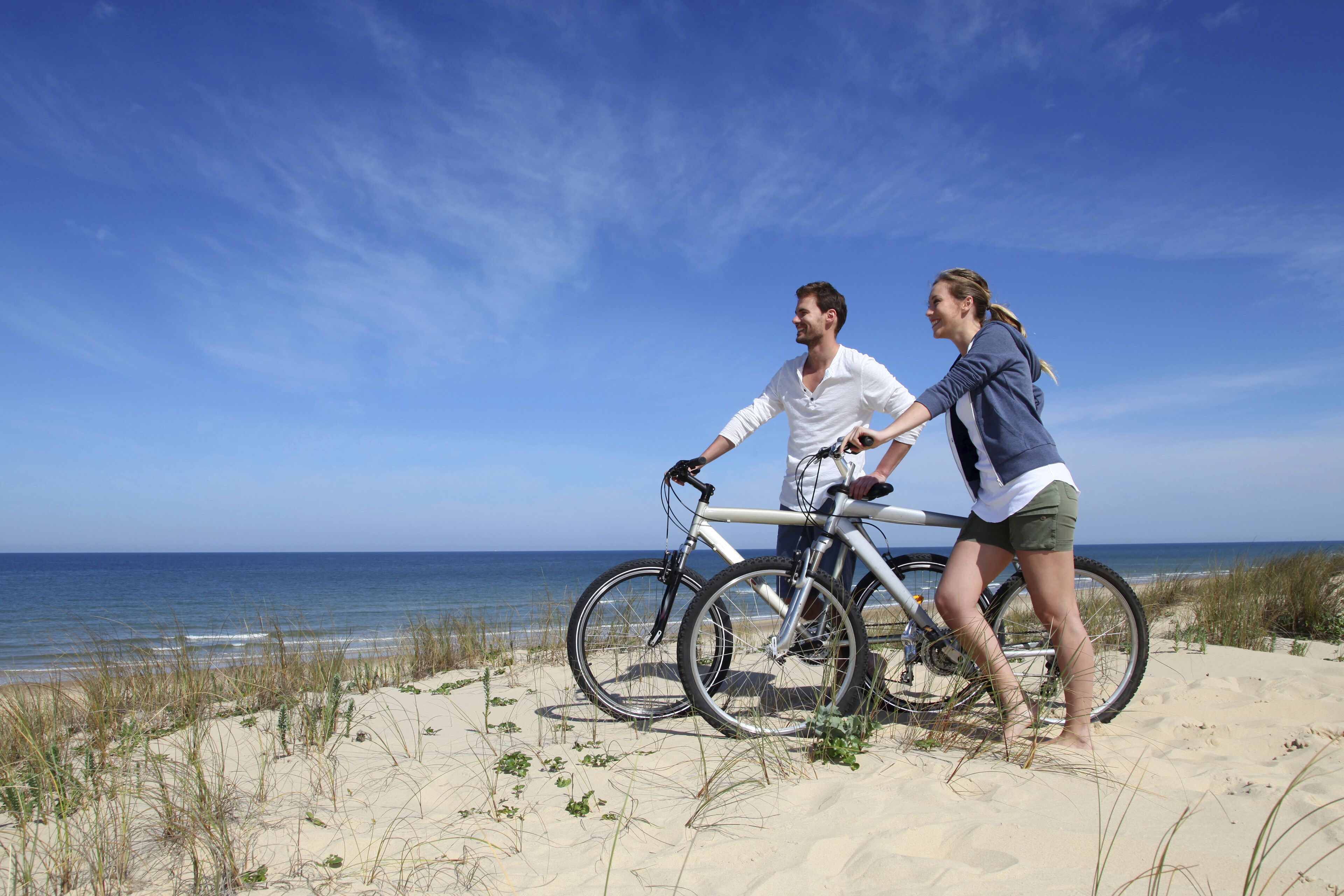 Escena de playa con dos personas en bicicletas, una sosteniendo a la otra, cerca del océano.