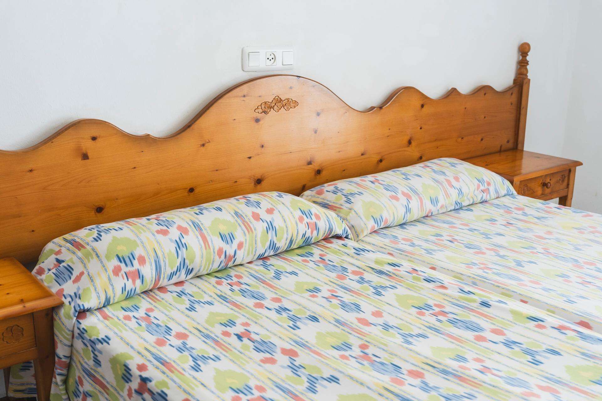 A wooden bed with a floral-patterned bedspread and matching pillows, a wooden nightstand, and a wall socket above the bed.