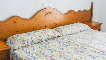 Una cama de madera con colcha y almohadas estampadas con motivos florales, una mesita de noche de madera, y un enchufe sobre la cama.