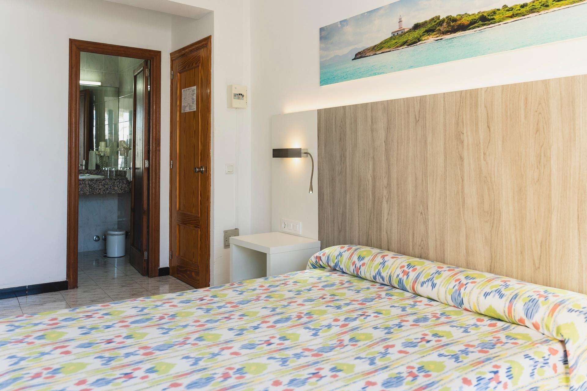 Un dormitorio con cama, cabecera de madera, baño con espejo y un cuadro de una escena costera.