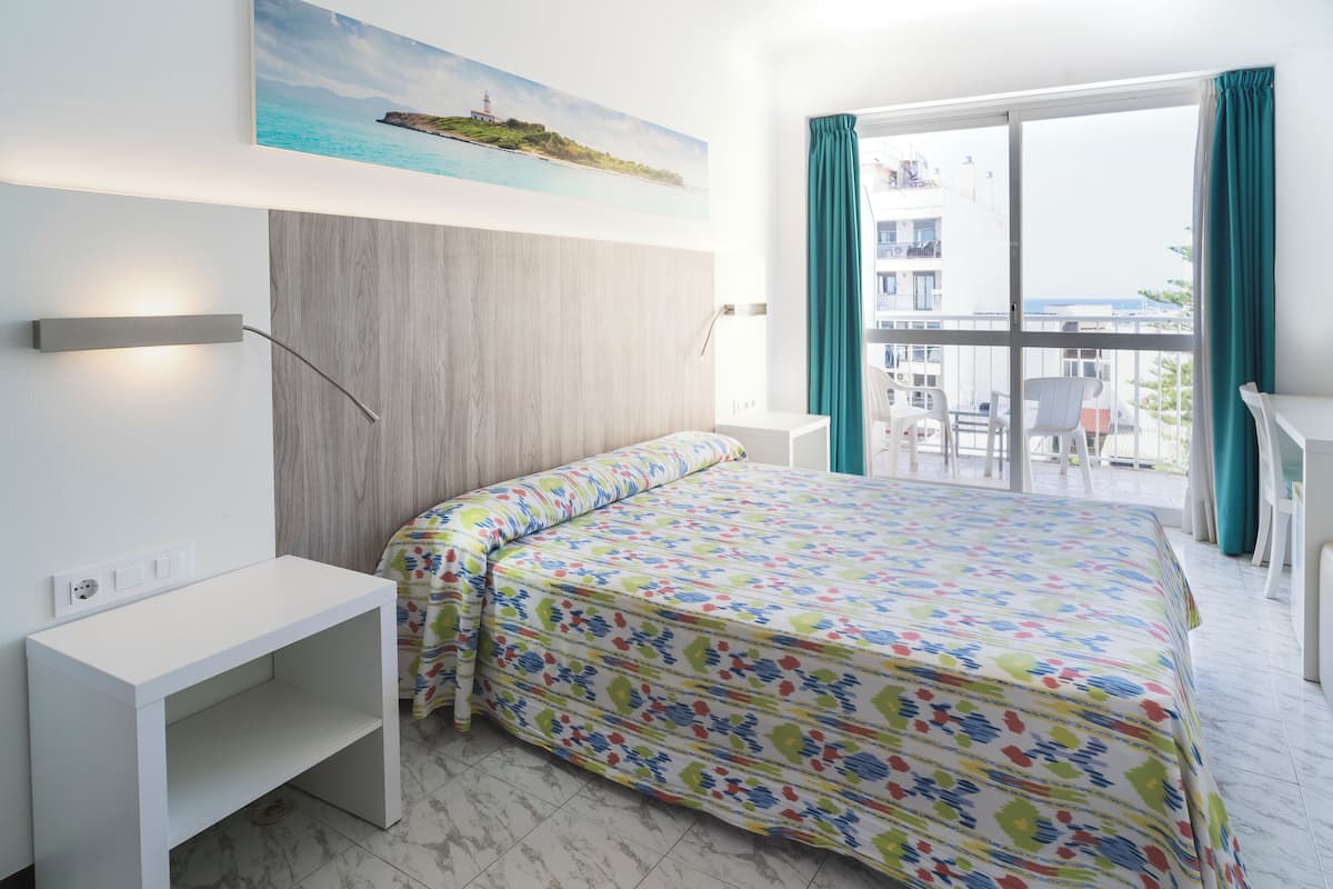 Een slaapkamer met een bed, een wit bureau, een wandlamp, een schilderij en een balkon met uitzicht.