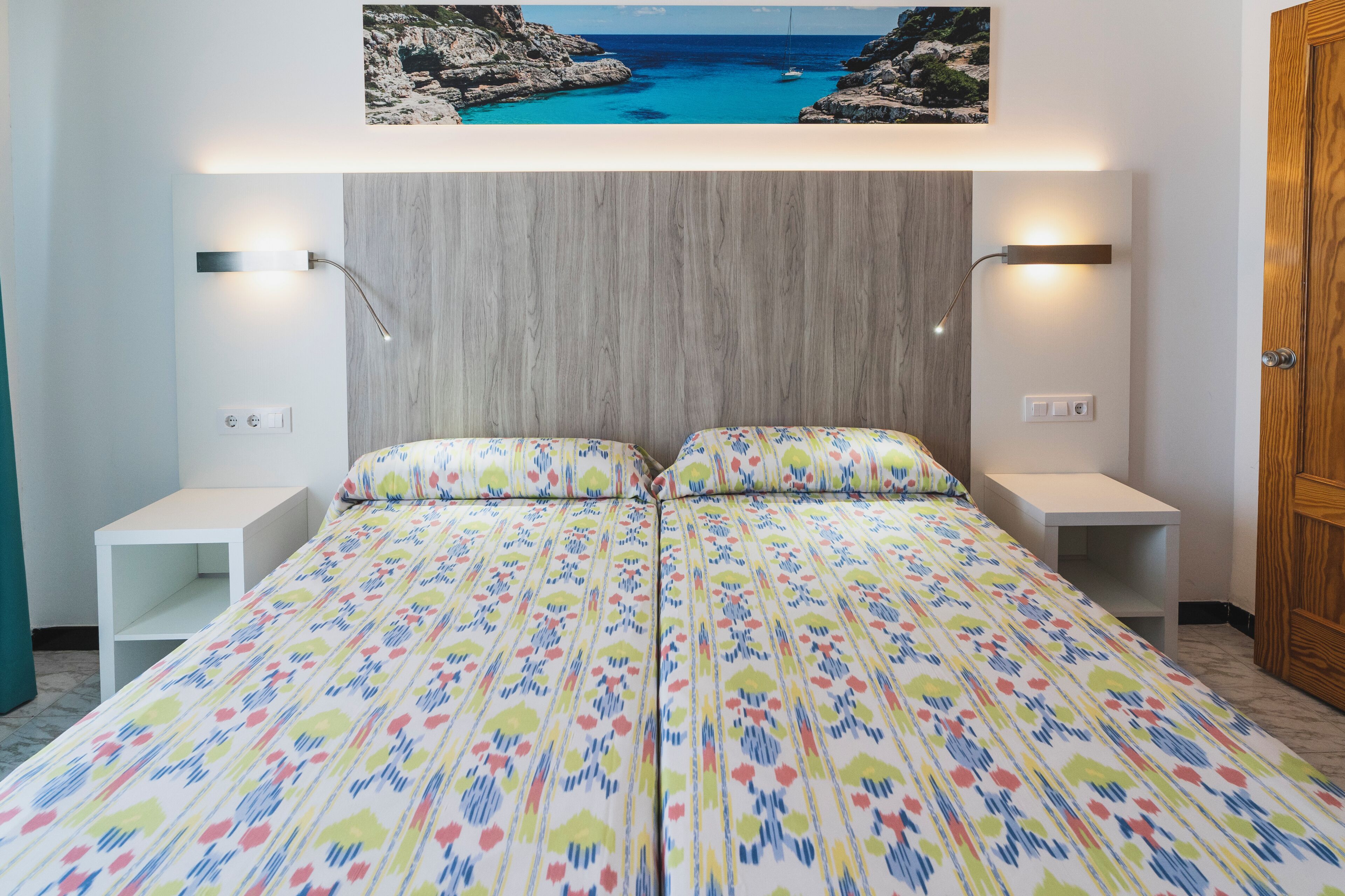 Een tweepersoonsbed met een kleurrijke bloemenbedekking, twee nachtkastjes, een houten hoofdbord en een ingelijst beeld boven het bed.