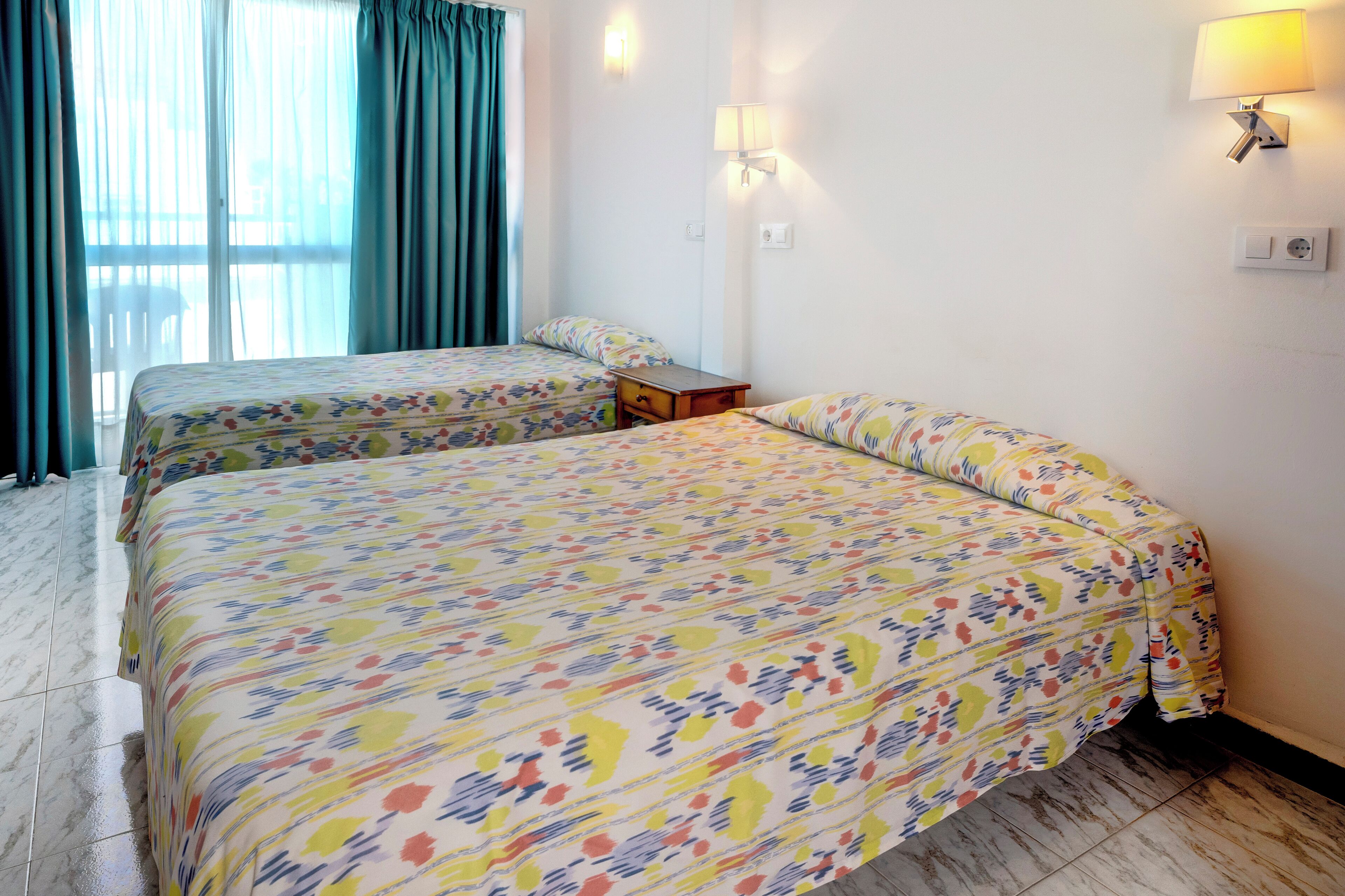 Een hotelkamer met twee bedden, een kleine tafel, een lamp en een raam met gordijnen.