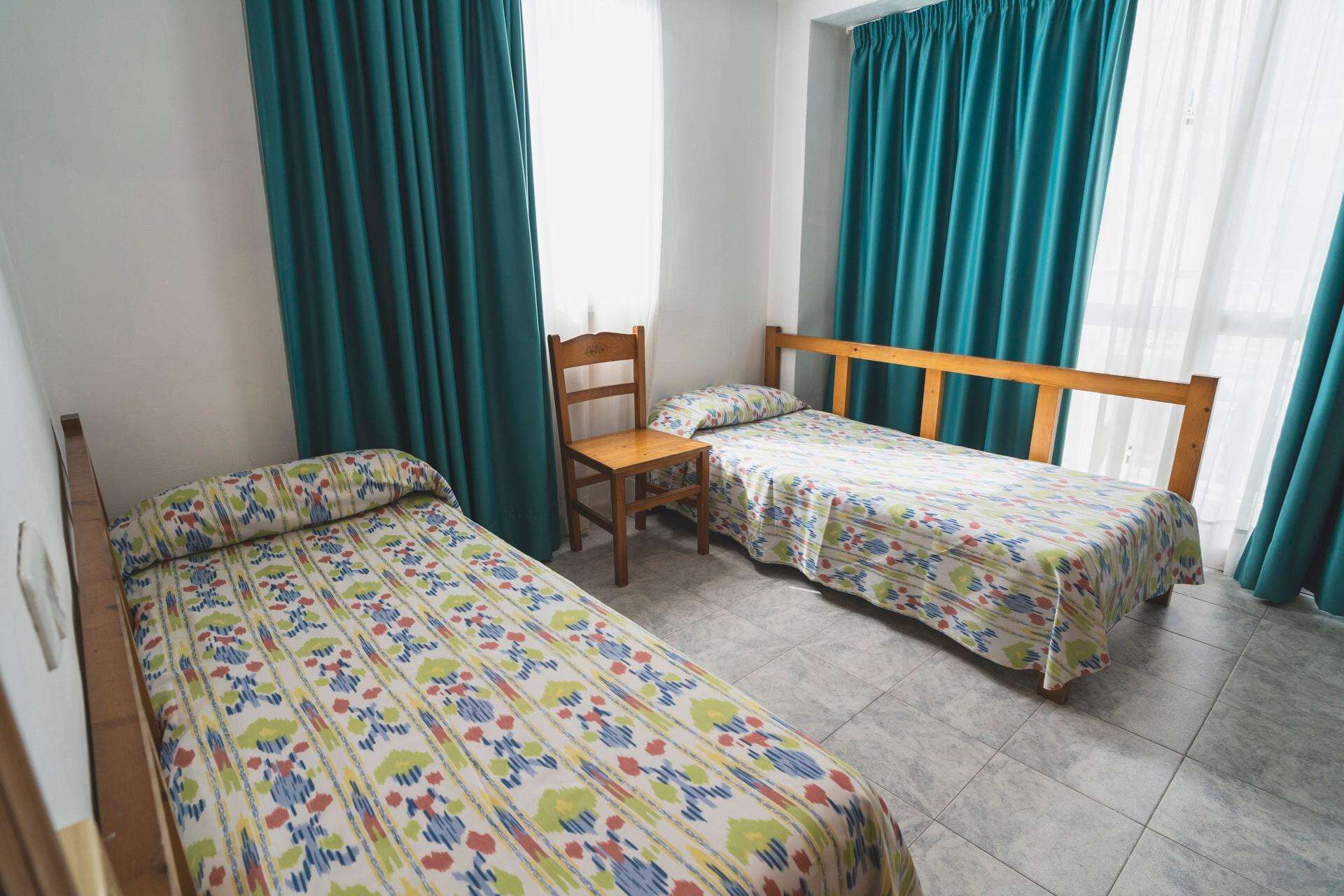 A room with two single beds, a wooden chair, and a window with curtains.